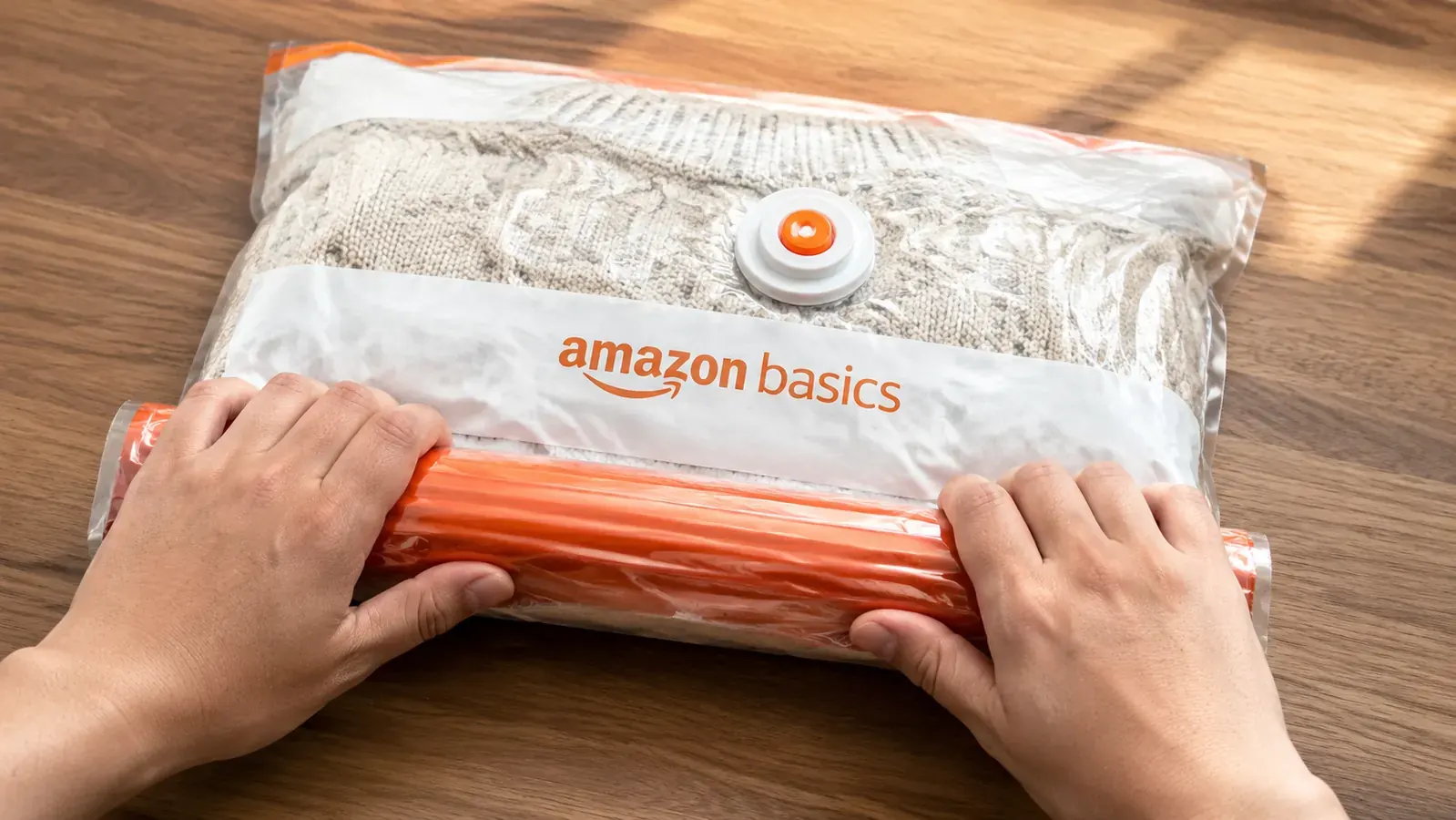 Hands rolling air out of a vacuum storage bag containing a folded down jacket on a wooden floor